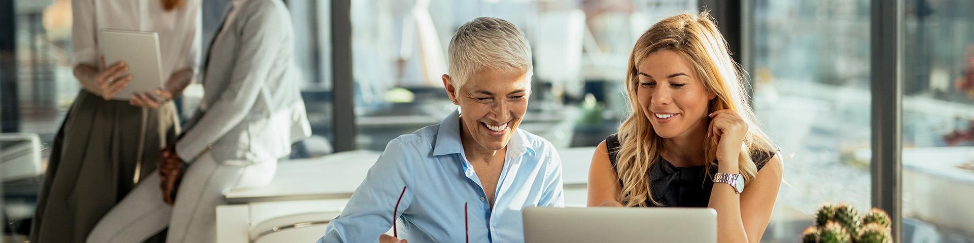 Two women sit together next to a bright window. They are looking at something on a laptop together. 