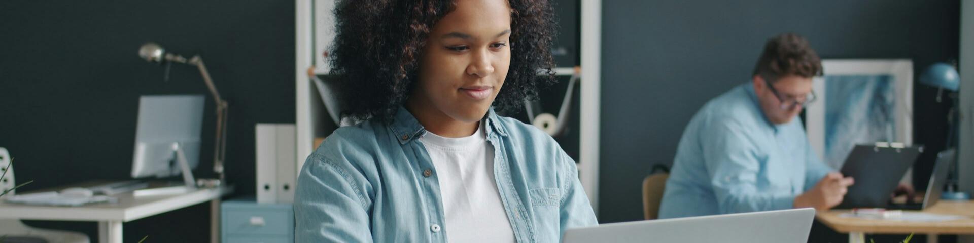 A person looking down at their laptop. Someone else works at a different desk in the background.